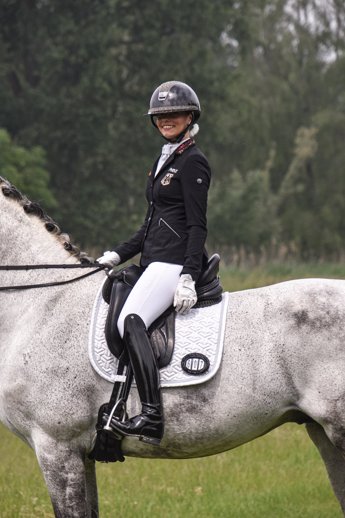 Smiling rider in a black jacket and crystal helmet on a grey horse, with a competition number on the saddle pad
