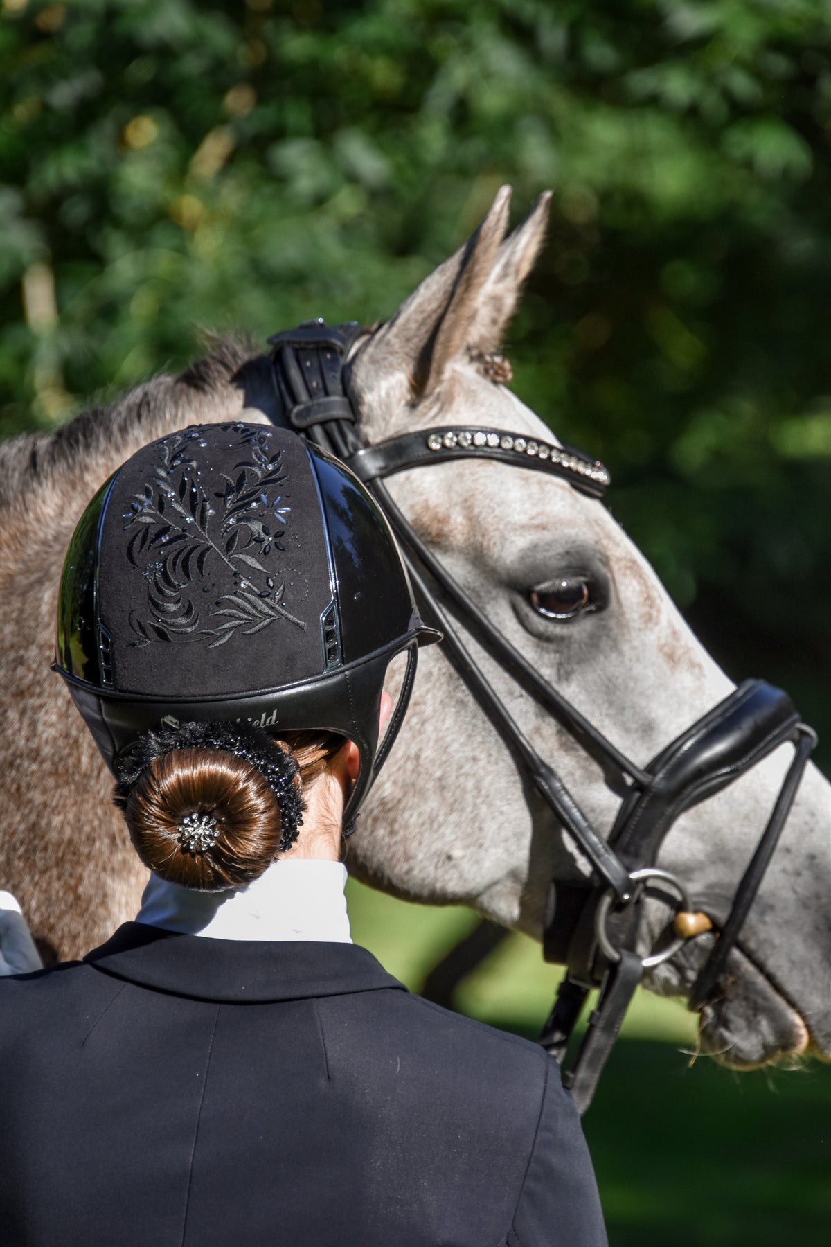 Reiterin von hinten mit schwarzem, besticktem Helm und glitzerndem BelliNoxx bun neben einem Apfelschimmel