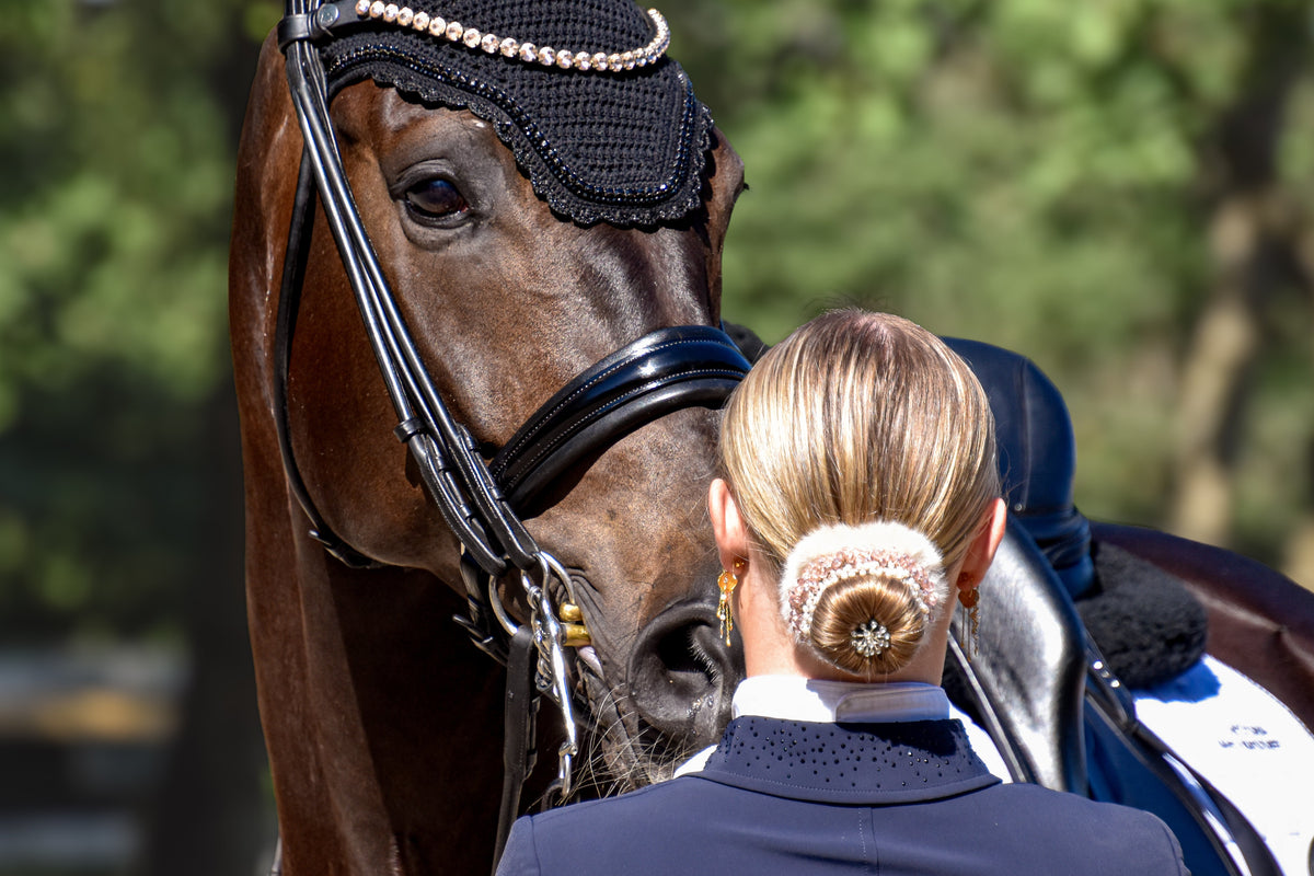 Rider seen from behind with a crystal-trimmed fur bun, beside a bay horse in a black ear bonnet