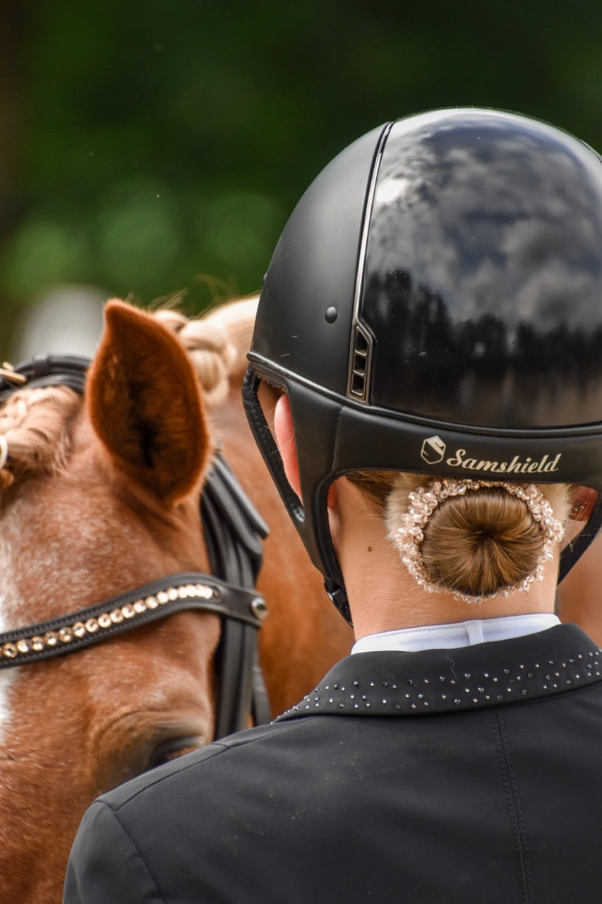 Rider seen from behind in a black helmet, with a crystal-trimmed bun just below the helmet, beside a chestnut horse